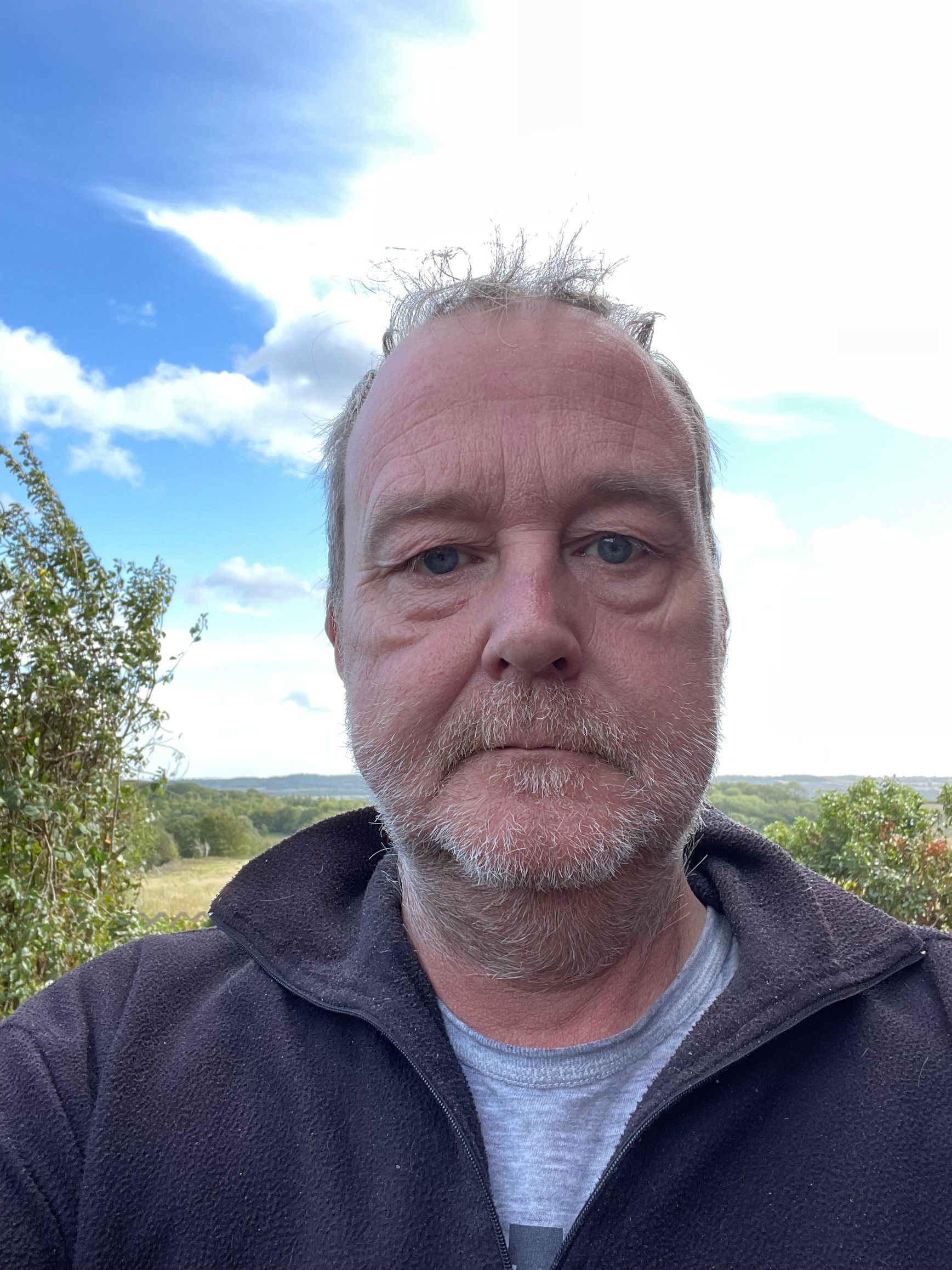 Man with beard against a scenic backdrop of trees and sky. He wears a grey t-shirt and dark jacket.