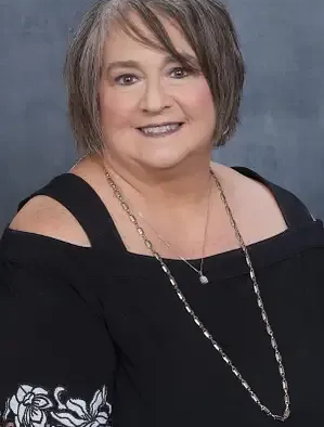 A person smiling in a portrait, wearing a black cold-shoulder top and a long silver chain necklace against a grey background.