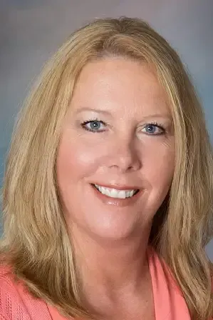 A person with blonde hair, blue eyes, and a pleasant smile, wearing a pink top against a neutral gray background.