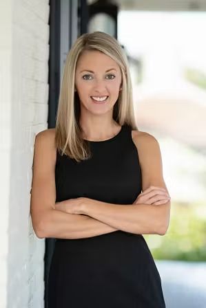 A person with shoulder-length blonde hair, wearing a black sleeveless top, smiles while leaning against a white wall.