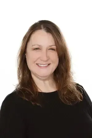 A person with long brown hair smiling against a plain white background, wearing a black long-sleeved shirt.