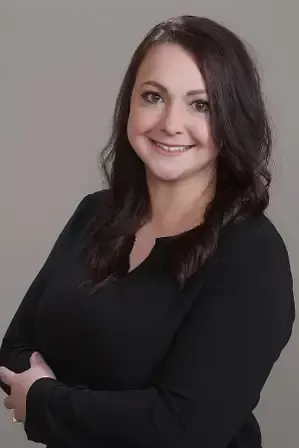 A person with dark hair smiling at the camera, wearing a black top, with their arms crossed, set against a gray background.