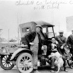 A black and white photo of a group of men standing next to an old car.