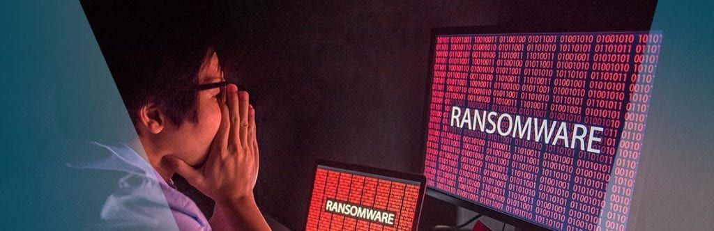 A man is sitting in front of a computer screen that says ' ransomware ' on it.