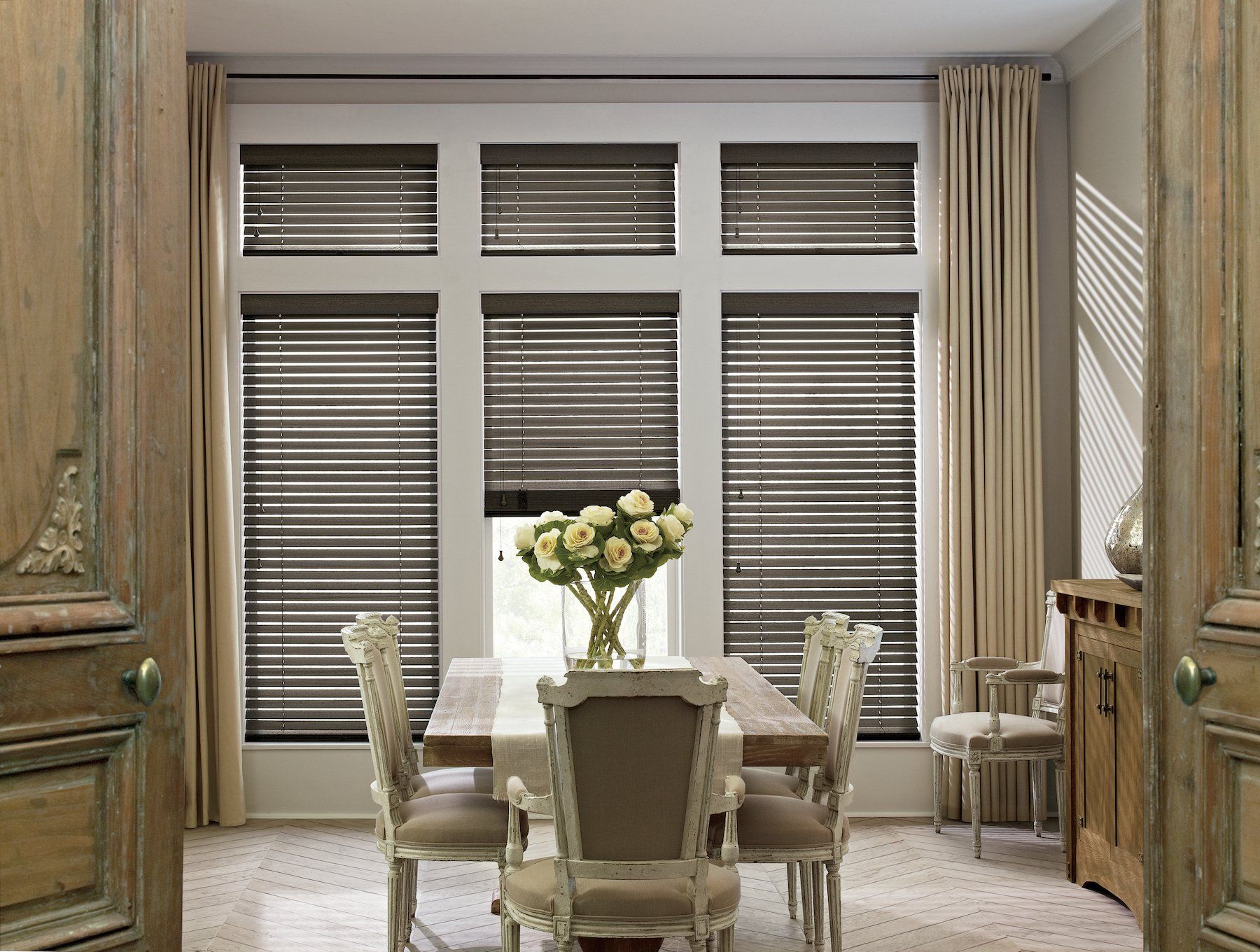 Dining room with wooden blinds, a table, chairs, and flowers in a vase. Light beige curtains.