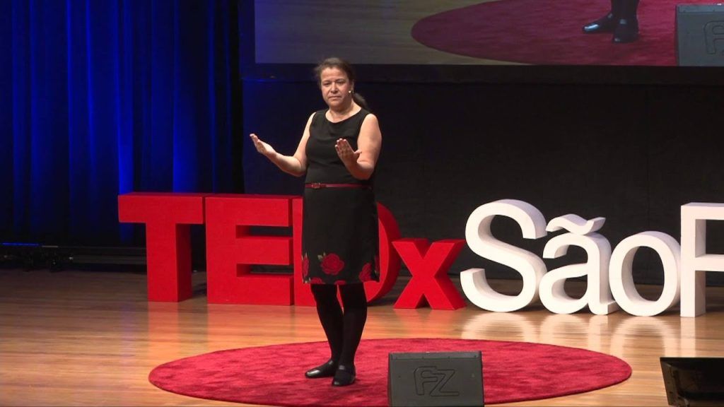 a mulher está de vestido preto, de pé, com as palmas das mãos viradas para cima. Ela está na frente de um letreiro do TedX São Paulo