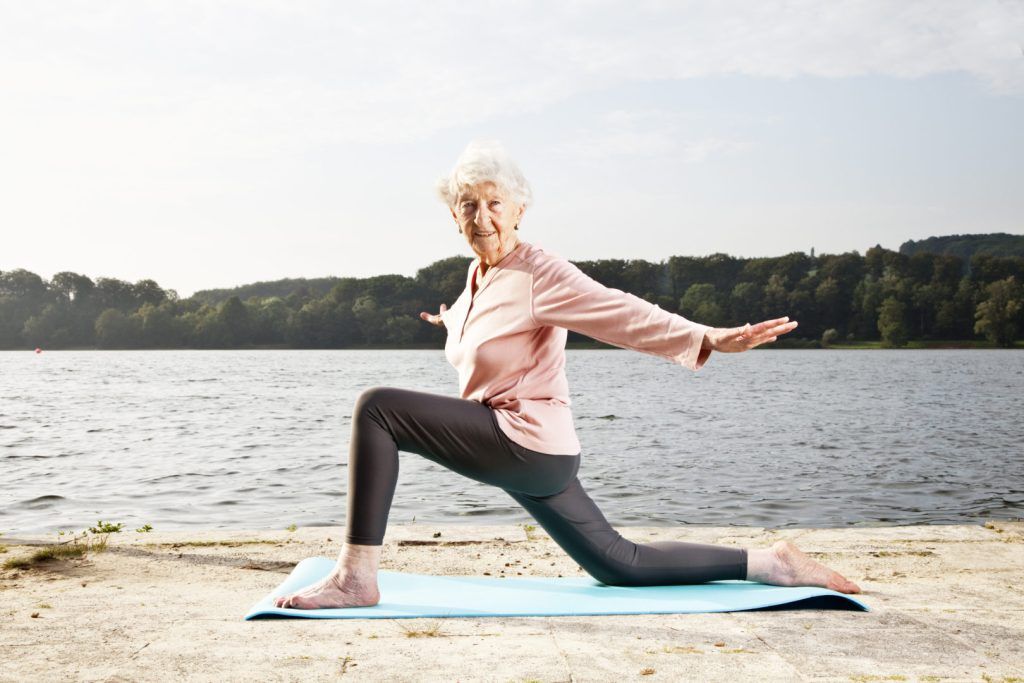 mulher madura fazendo ioga em frente ao lago. ela está em cima de um tapete de ioga azul.