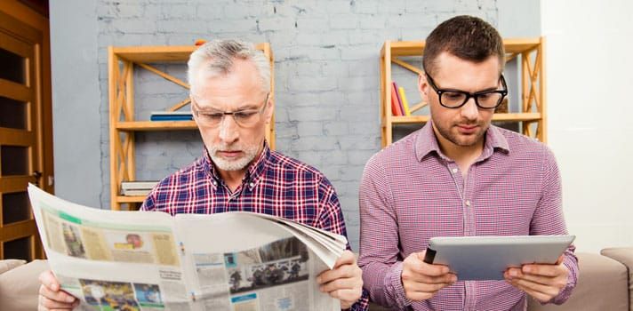 um maturi lê jornal enquanto ao seu lado um jovem lê as notícias em um tablet