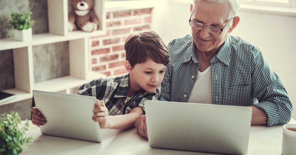 maturi e seu neto estão olhando para a tela de um computador. o menino olha atento enquanto seu avô sorri. os dois usam camisa de botão xadrez.