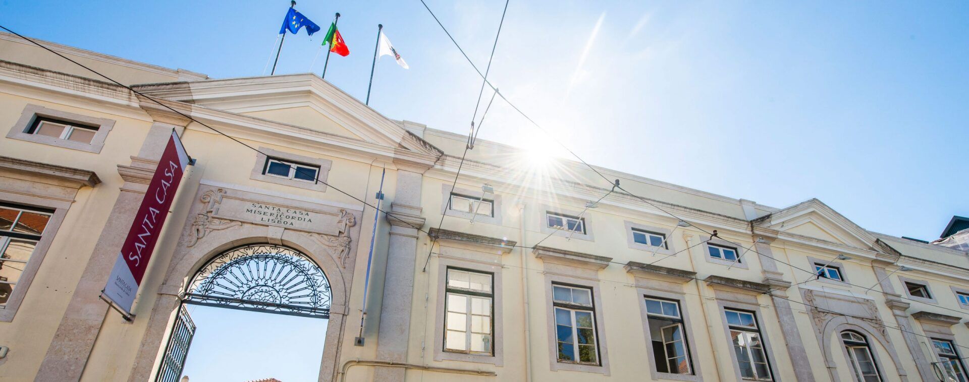foto da faixada Santa Casa de Misericórdia de Lisboa em Portugal. O céu está azul e o sol brilha, em cima da construção pode-se ver três bandeiras.