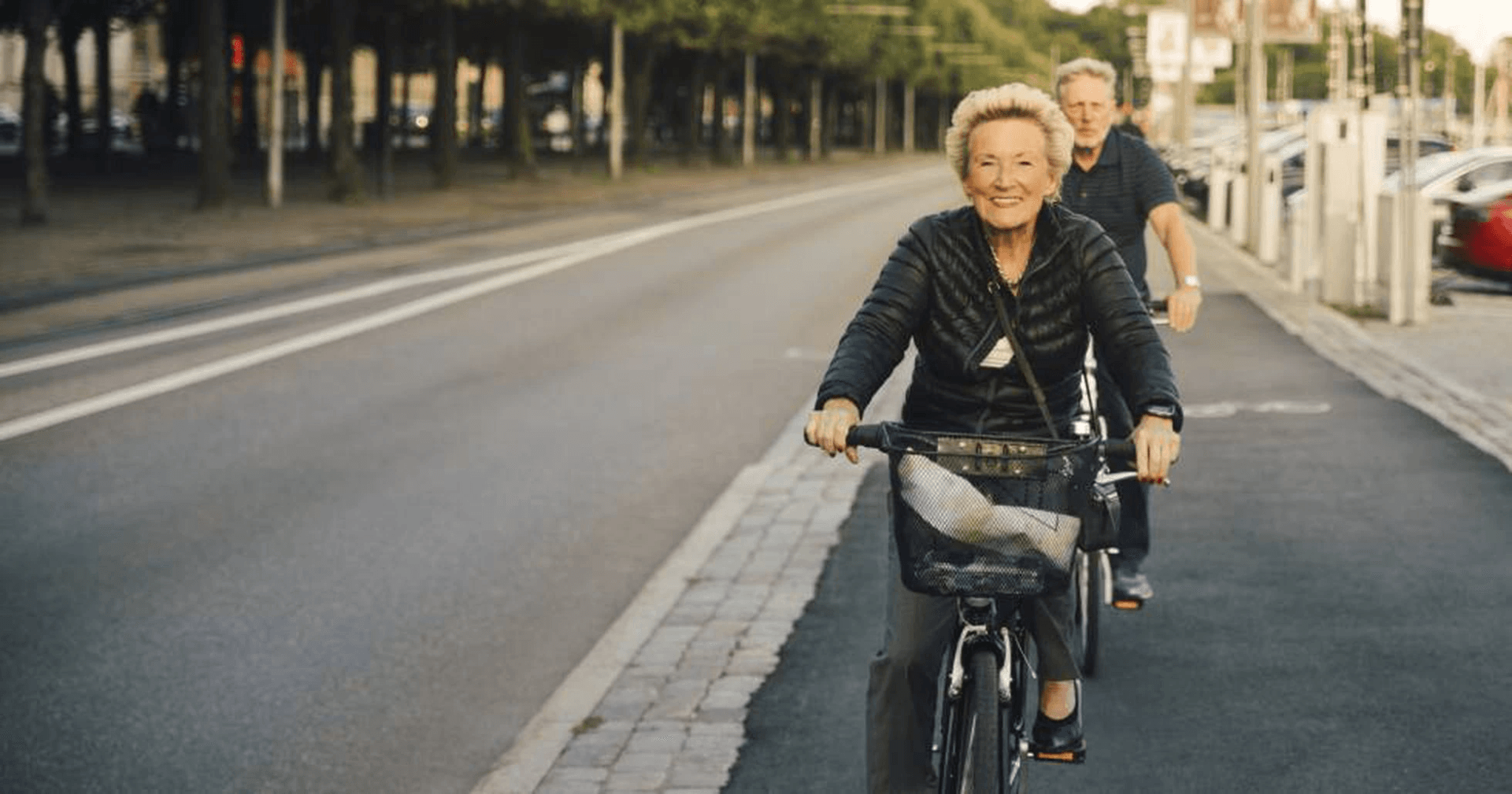 maturis andando de bicicleta felizes. a mulher está de preto e segue com a bicicleta na frente. o homem, que vai atrás, também está de preto.