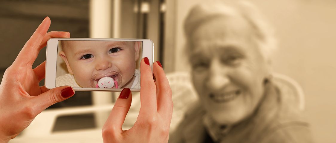 na imagem aparece uma mulher tirando uma foto de uma senhora madura, porém, na tela do celular aparece a foto de um bebê com chupeta na boca