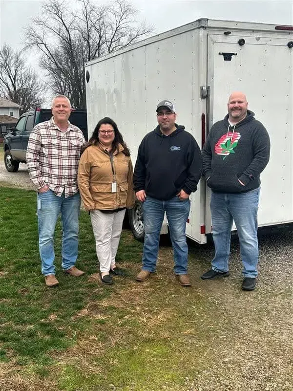 Four people standing outdoors beside a white trailer on a grassy yard, wearing casual clothes.