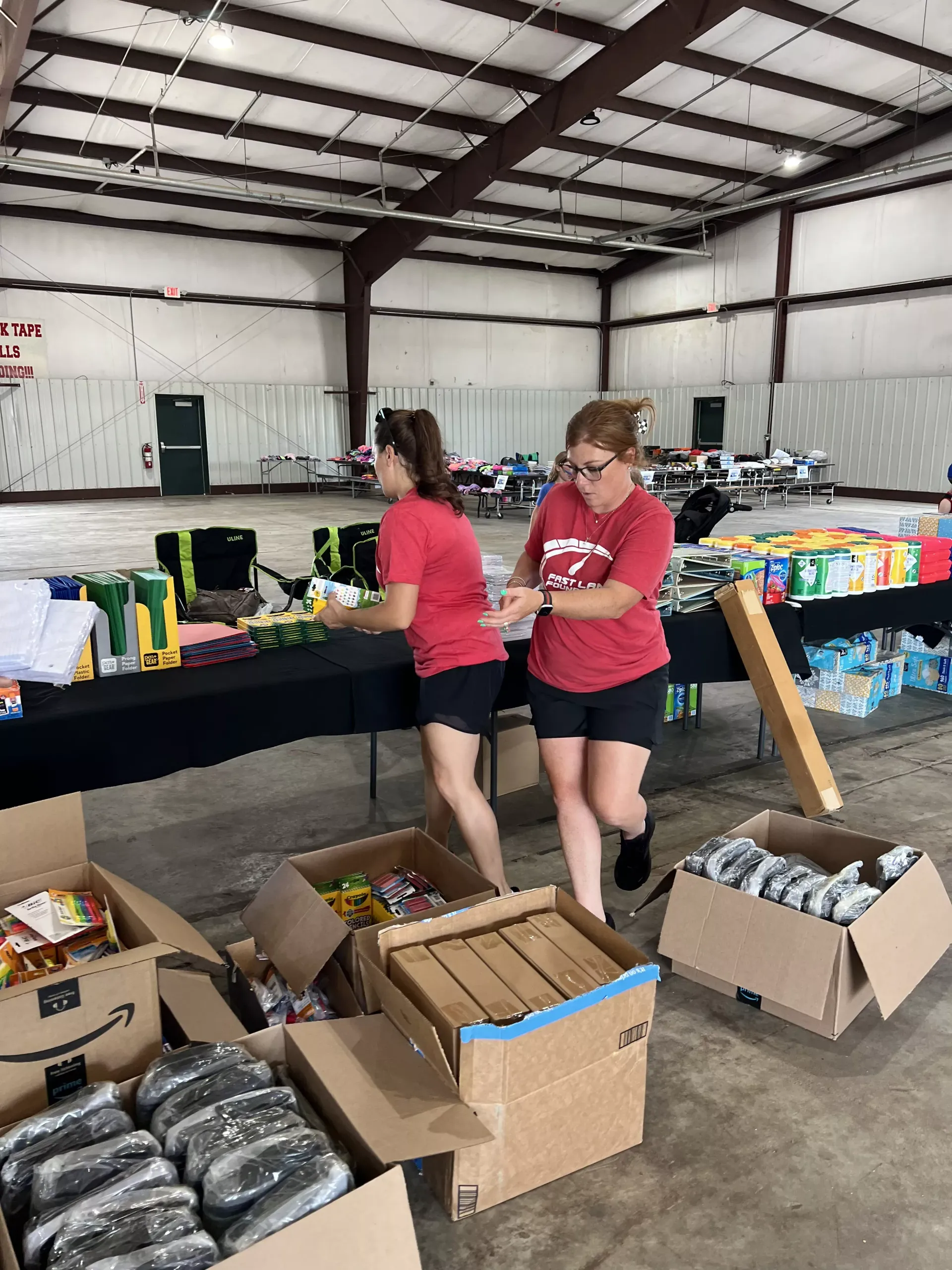 Two volunteers in red shirts sort donations at a table in a warehouse, with boxes and supplies stacked around them.
