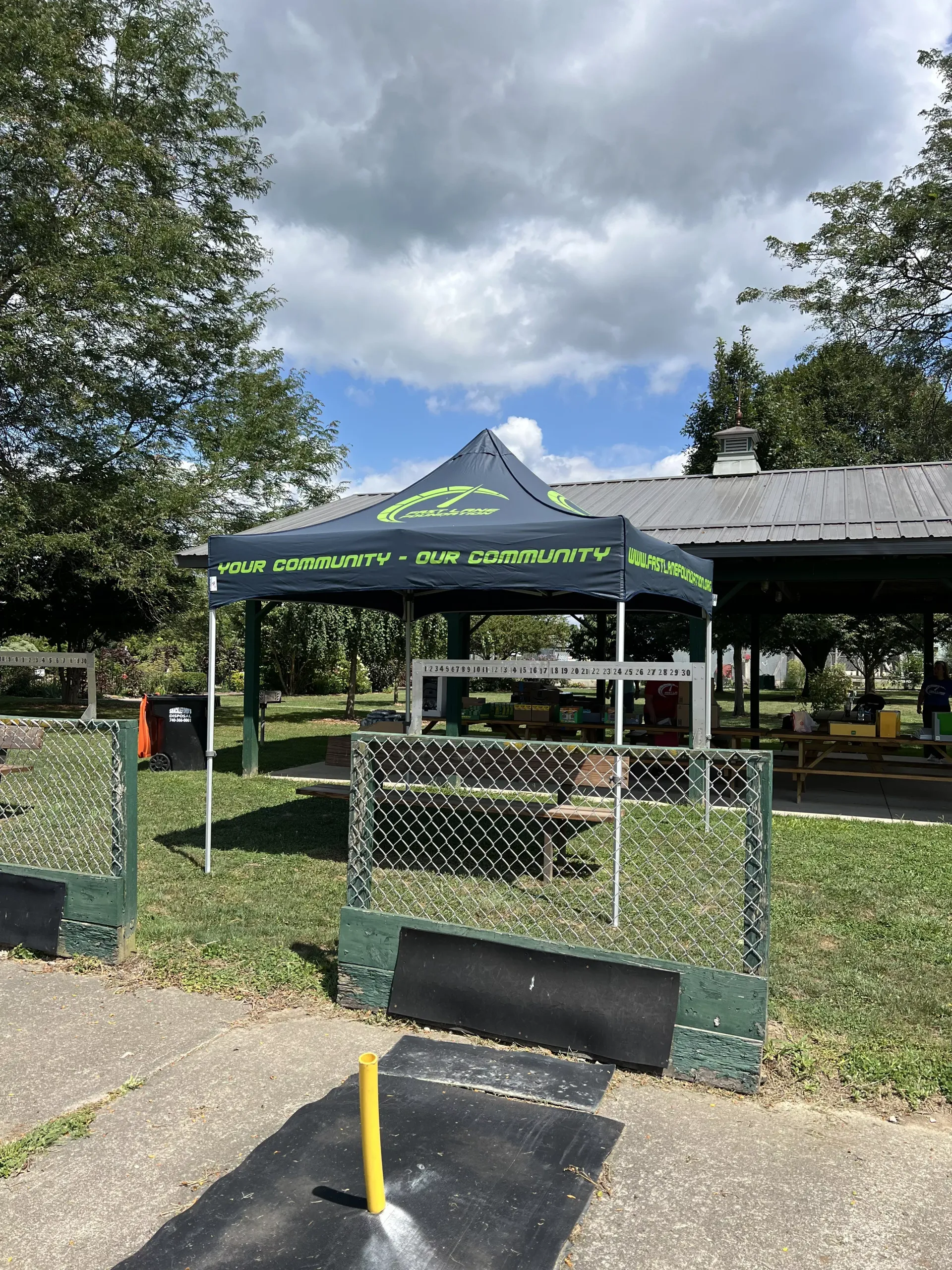 Outdoor tented checkpoint with chain-link fence, black mat, and yellow post beside a park shelter under cloudy skies