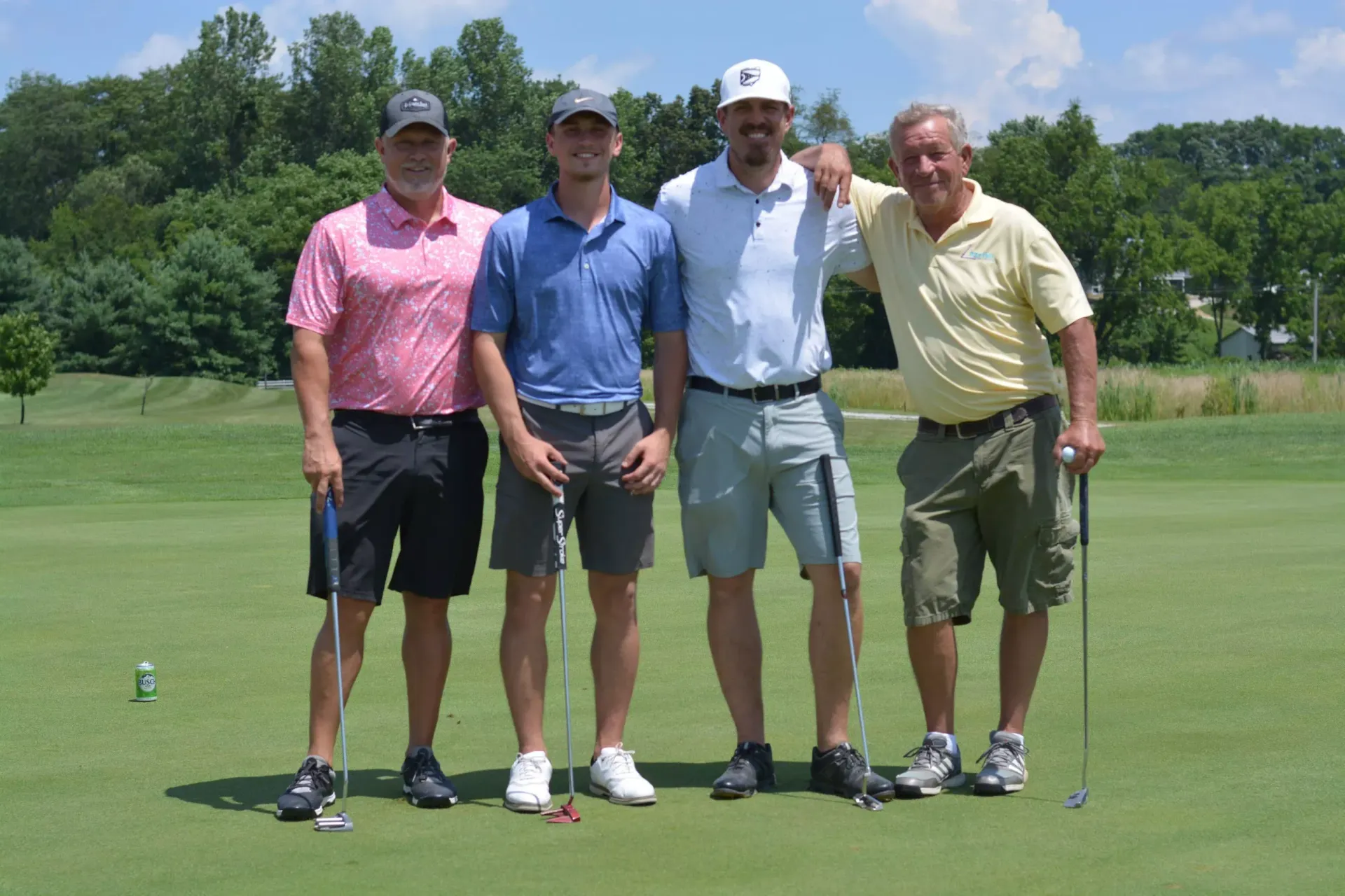 Four golfers standing together on a green golf course, holding clubs on a sunny day.