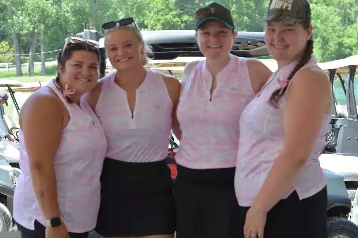 Four people in matching pink shirts and black skirts posing together outdoors, smiling at the camera.