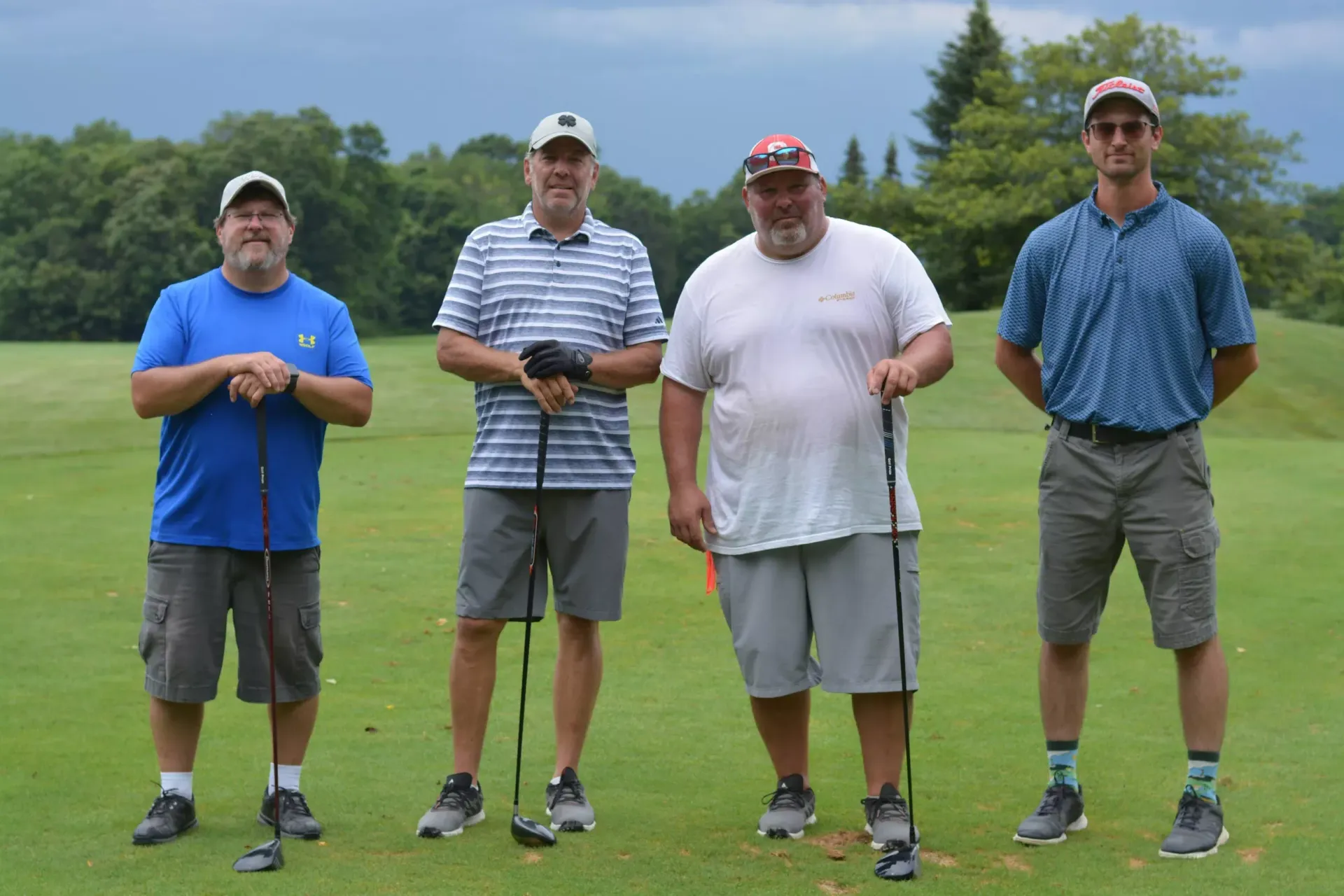 Four golfers standing on a tee box holding clubs on a grassy course under a cloudy sky.