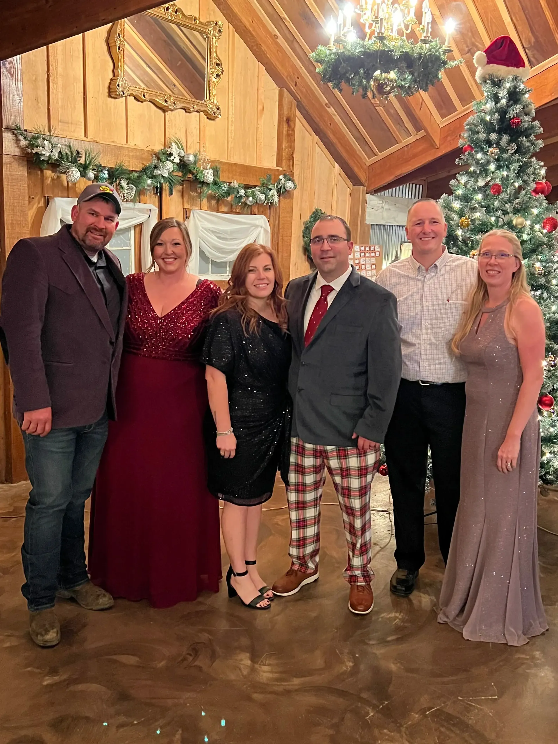 Six people posing together in a warmly lit, wood-paneled room decorated with a Christmas tree and chandelier.