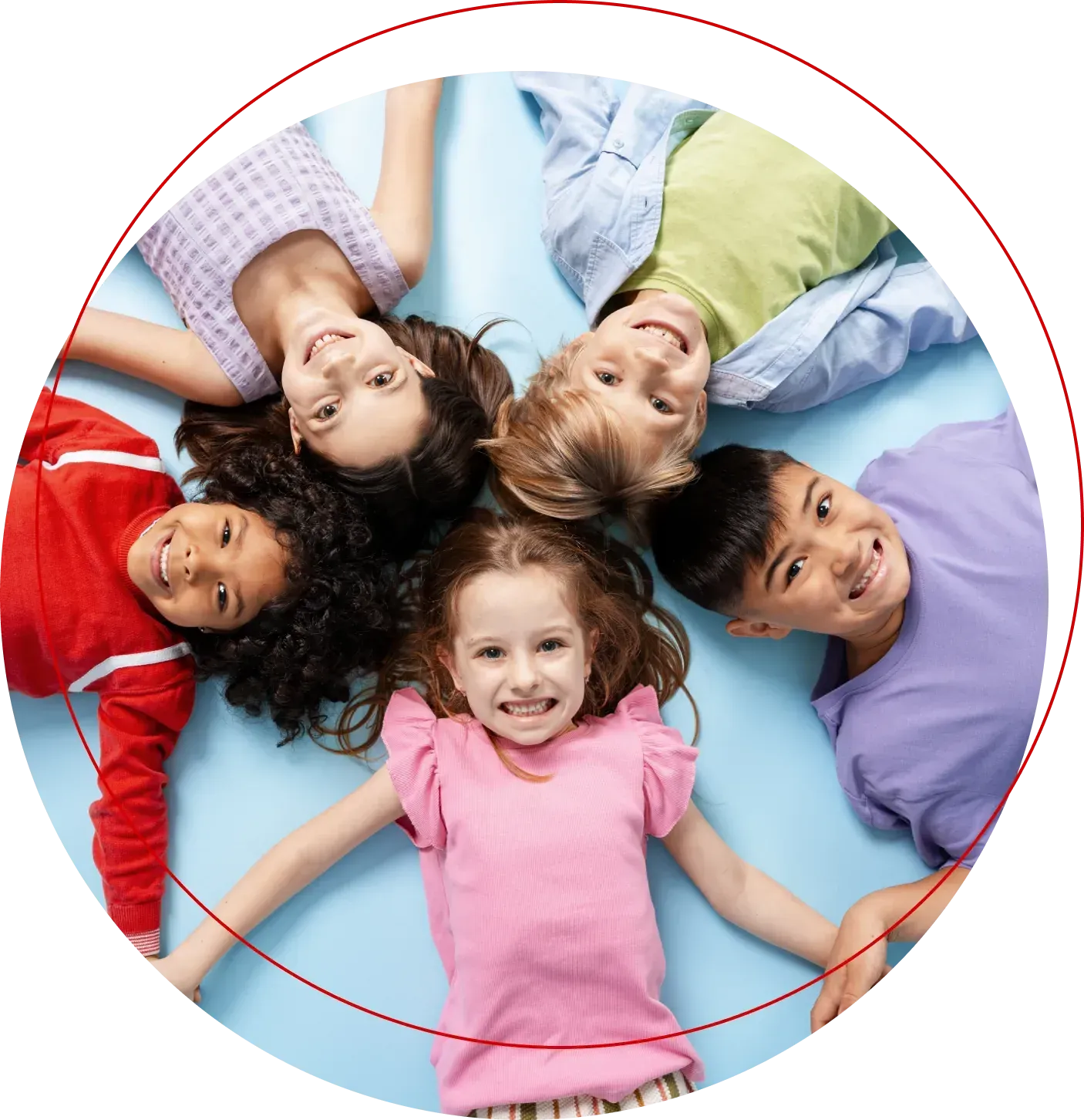 Five smiling children lying in a circle on the floor, heads together, looking up at the camera.