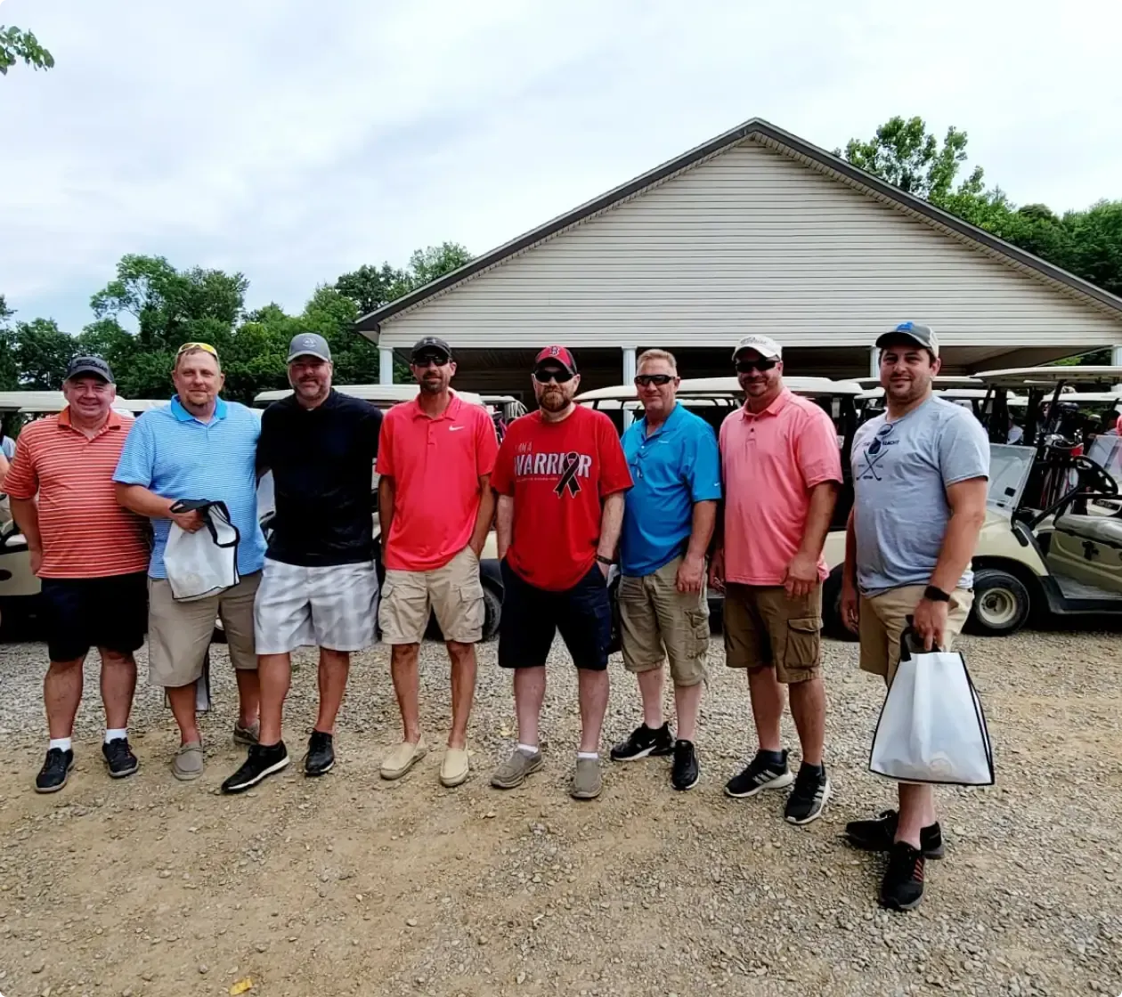 Eight men stand together on a gravel lot in front of a beige building, posing for a group photo.