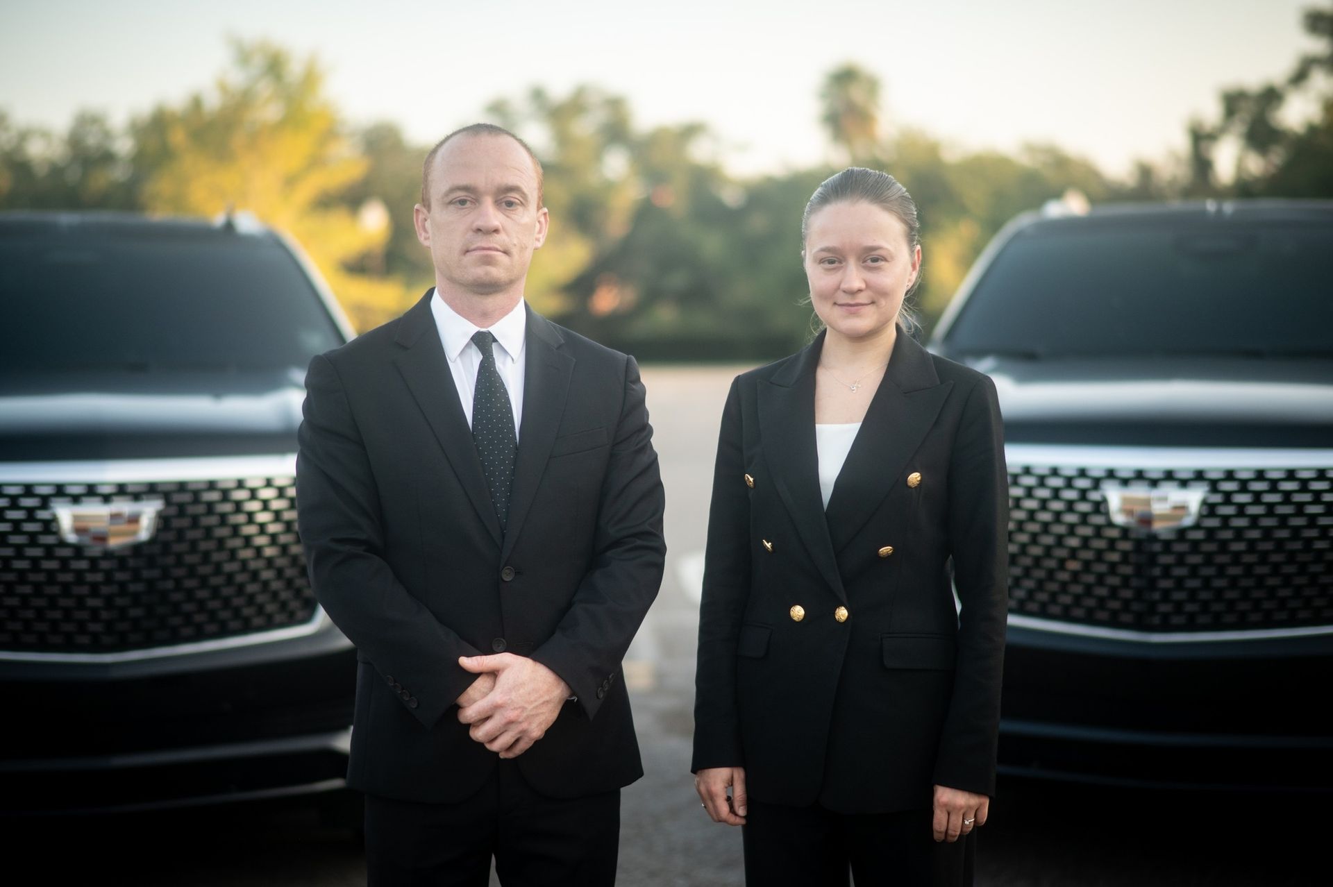 Two people in black suits stand in front of two black Cadillac Escalades.