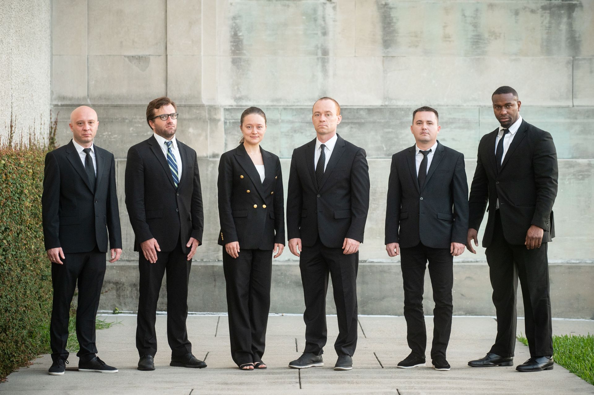 Two people in black suits stand in front of two black Cadillac Escalades.