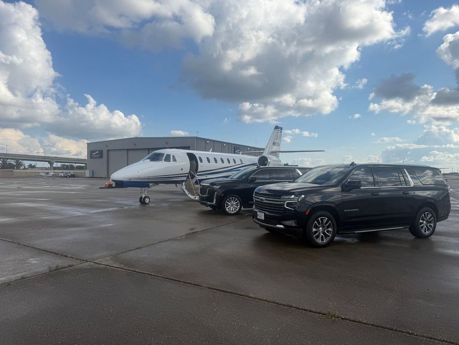 Black SUV parked beside a private jet on a tarmac at an airport. Clear blue sky.