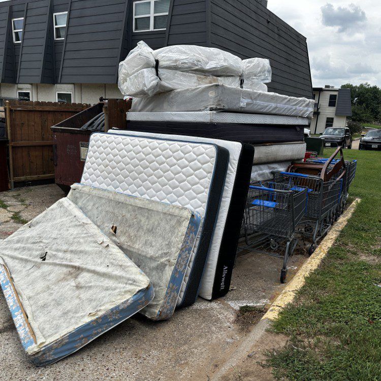 Pile of discarded mattresses and furniture next to a dumpster and shopping cart, in front of an apartment building.