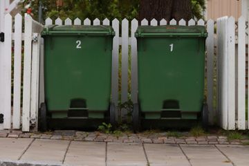 Two green trash cans with numbers 1 and 2, behind a white picket fence on a brick path.