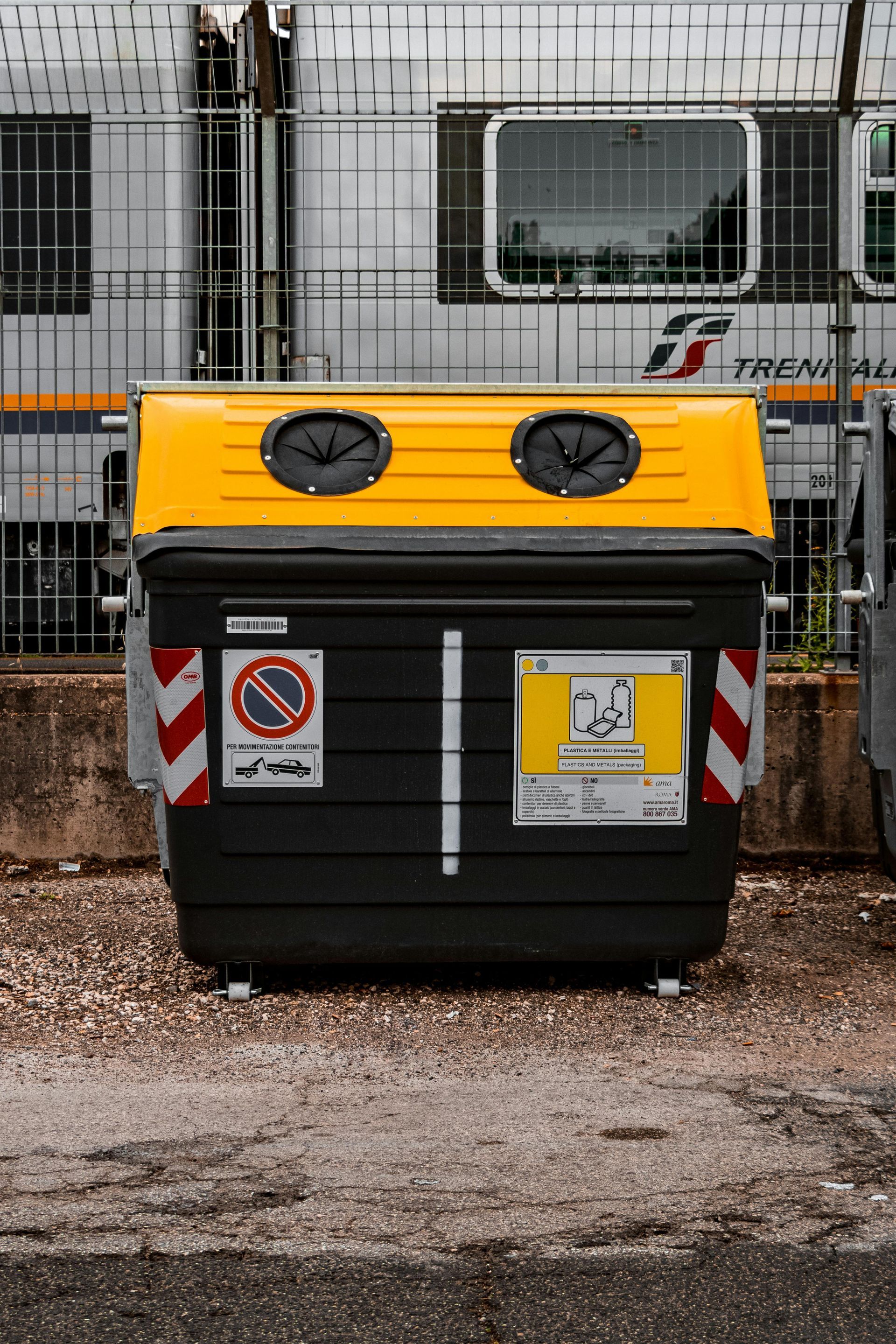Yellow and black recycling bin at a train station, with a train in the background.