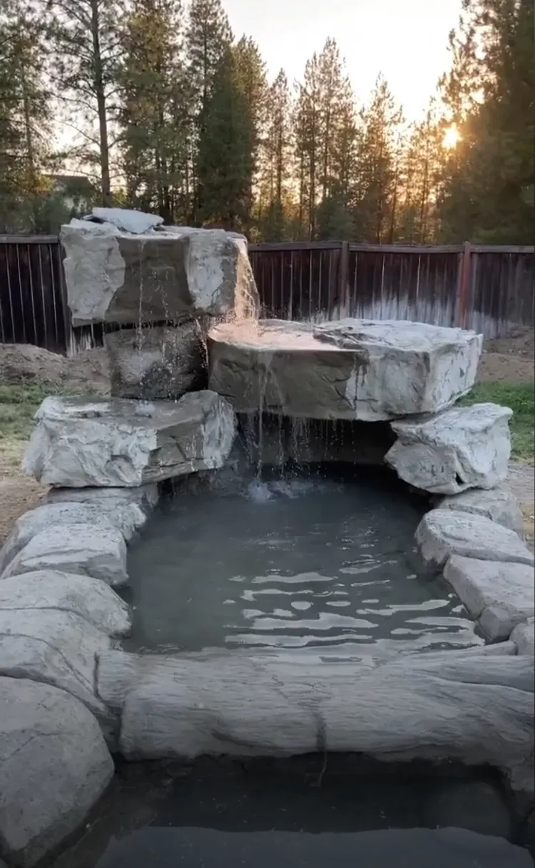 Stone waterfall feature with cascading water into a pond, surrounded by faux rocks. Sunset in background.