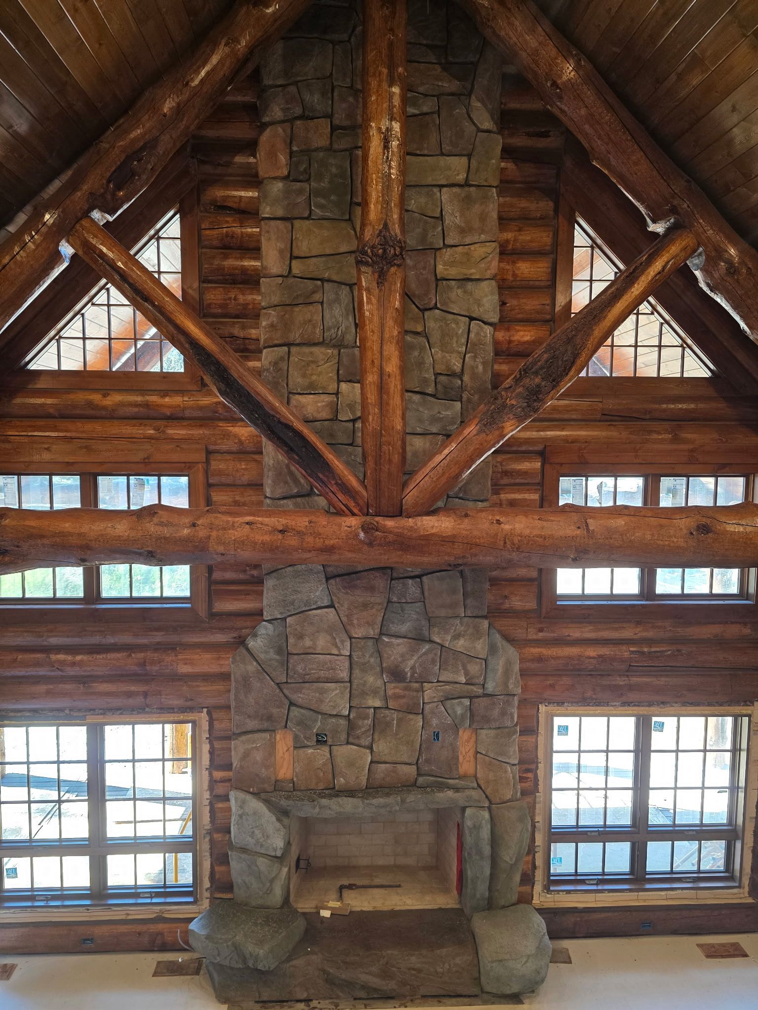Stone fireplace in a rustic cabin, flanked by windows. Wooden beams and ceiling.