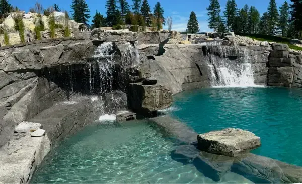 Waterfalls cascading into a turquoise pool, surrounded by gray rock formations and trees under a blue sky.