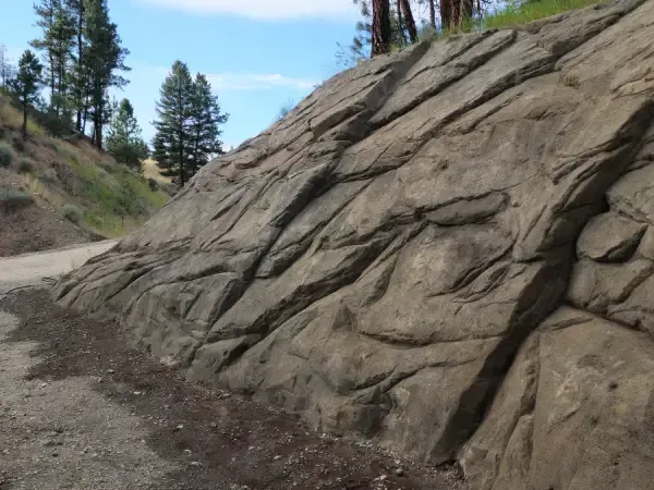 Rock wall alongside a dirt road in a natural setting; trees and blue sky visible.