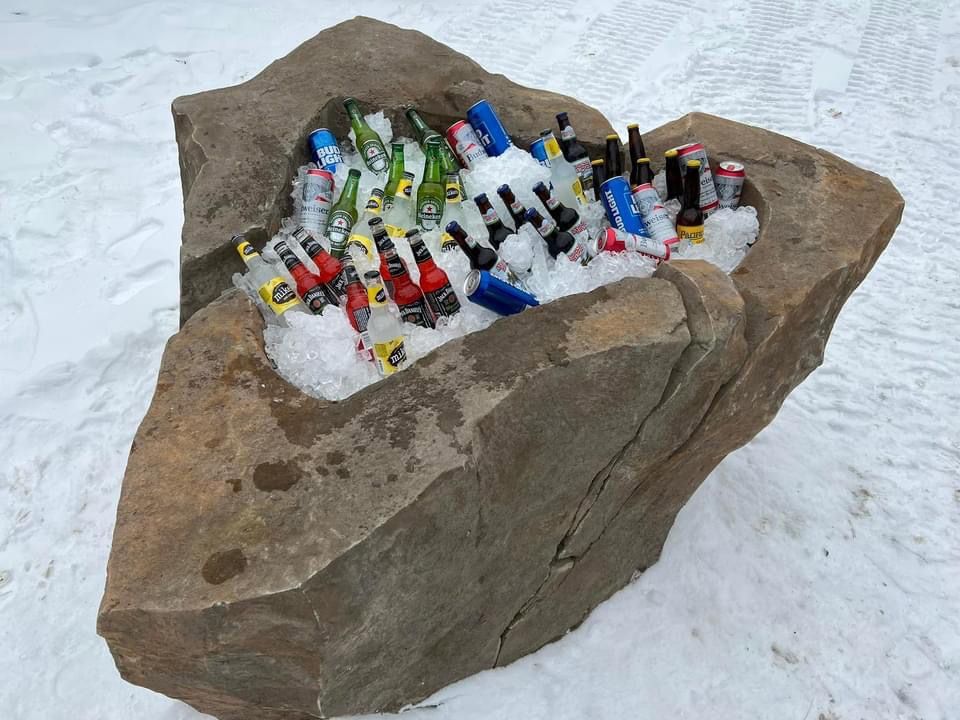 Rock cooler filled with ice and assorted bottled beverages, set in snowy outdoor environment.