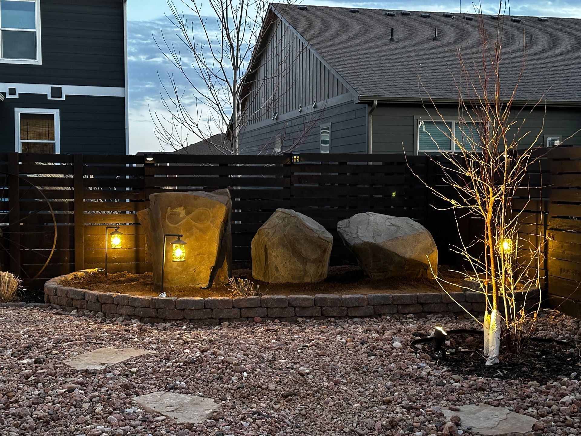 Backyard rock garden with dark fence, lit by solar lights. Rocks are center, tree right.