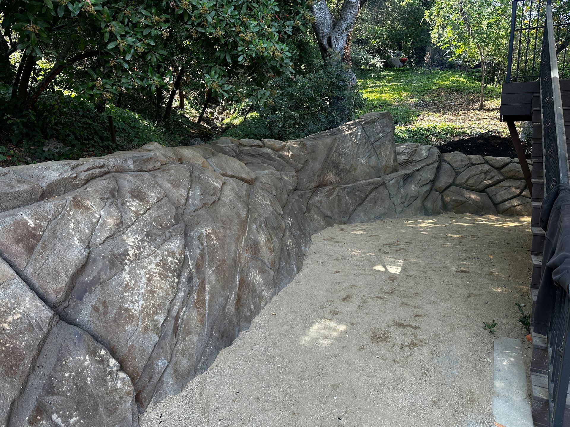 Rocky, stone-walled enclosure with sandy ground. Trees in the background.