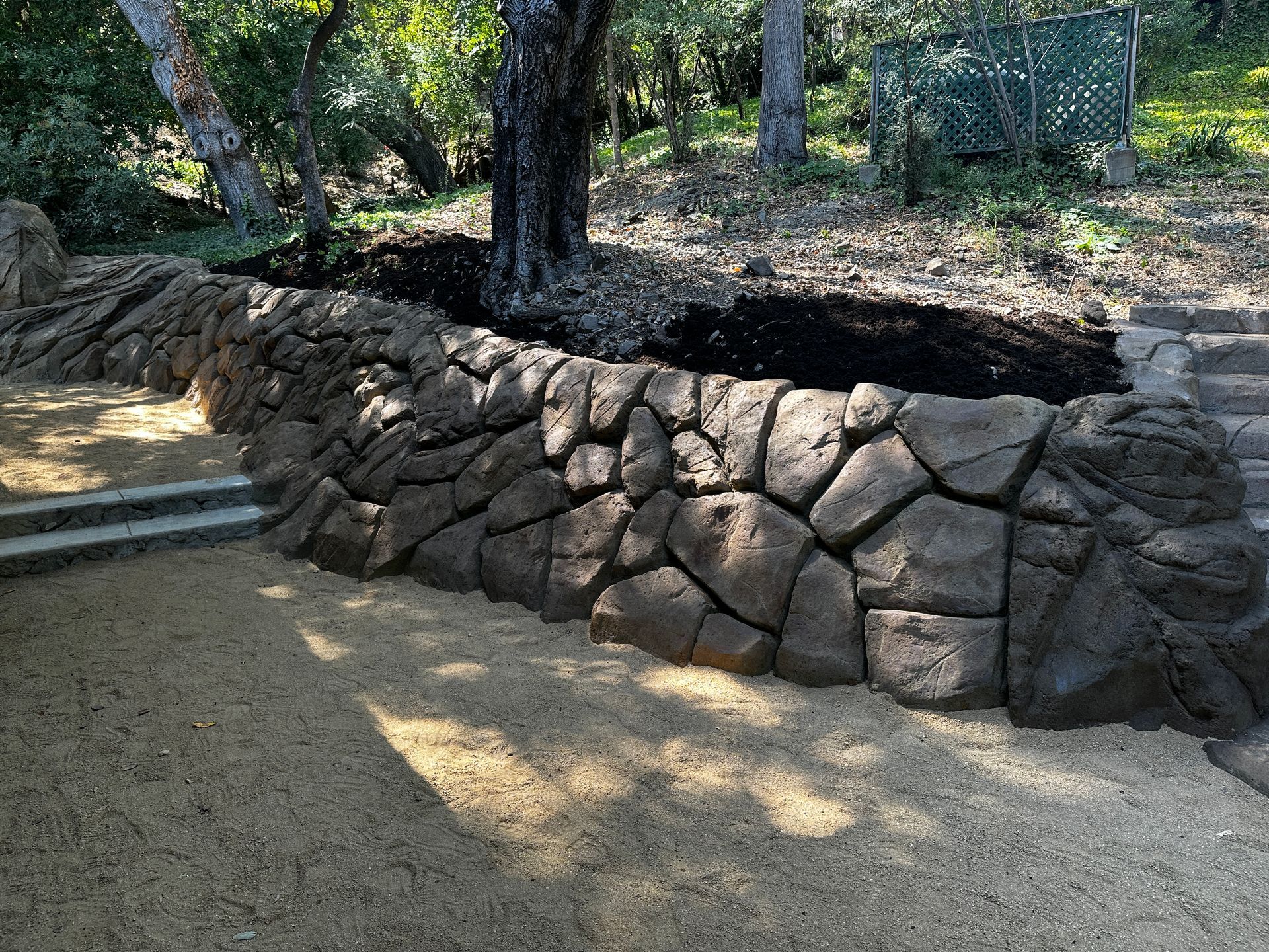 Stone retaining wall with steps, tree, and gravel ground in a yard.