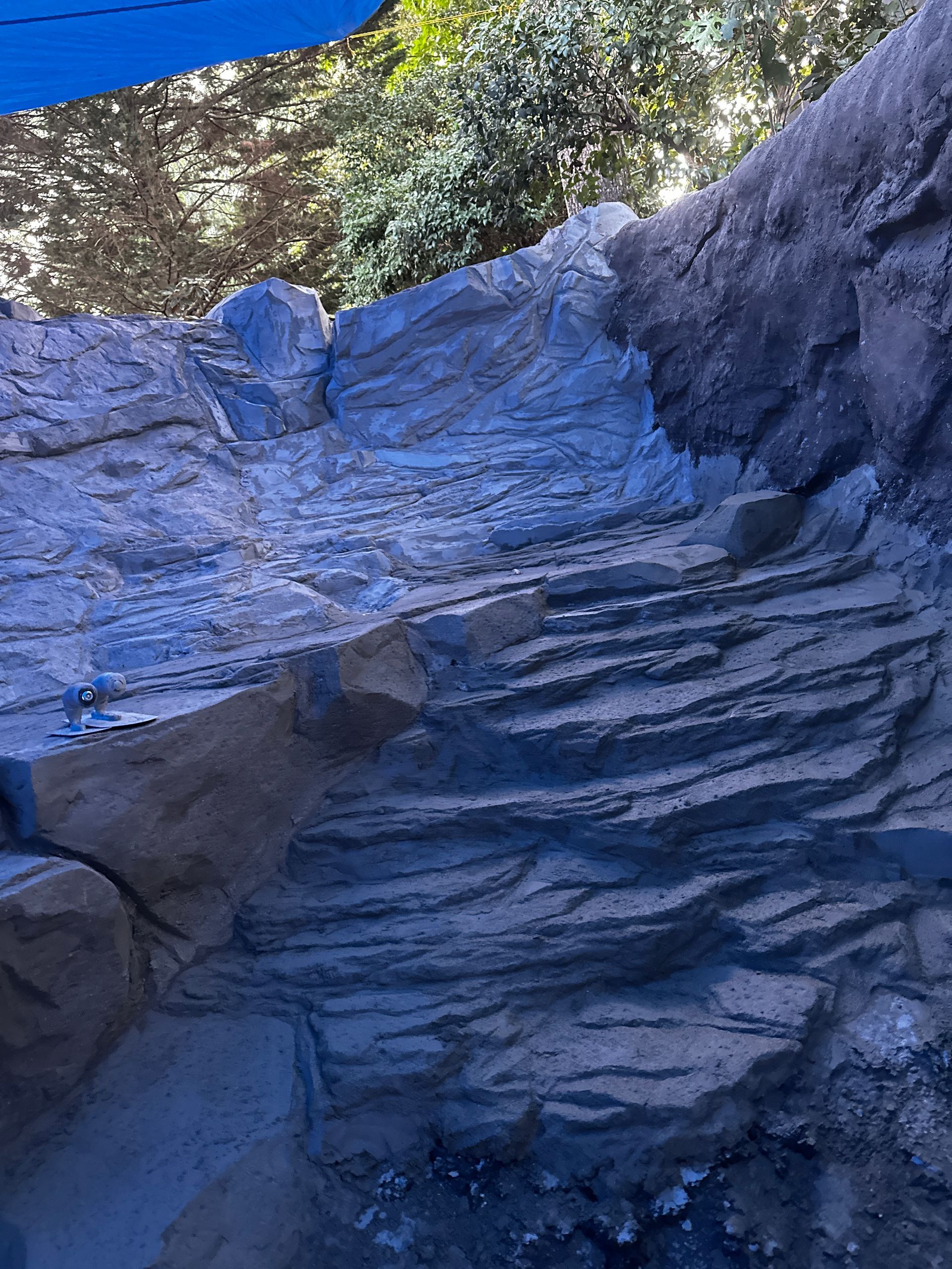 Rocky structure, possibly a water feature with layered, blue-gray and tan stone, shaded by a blue tarp.