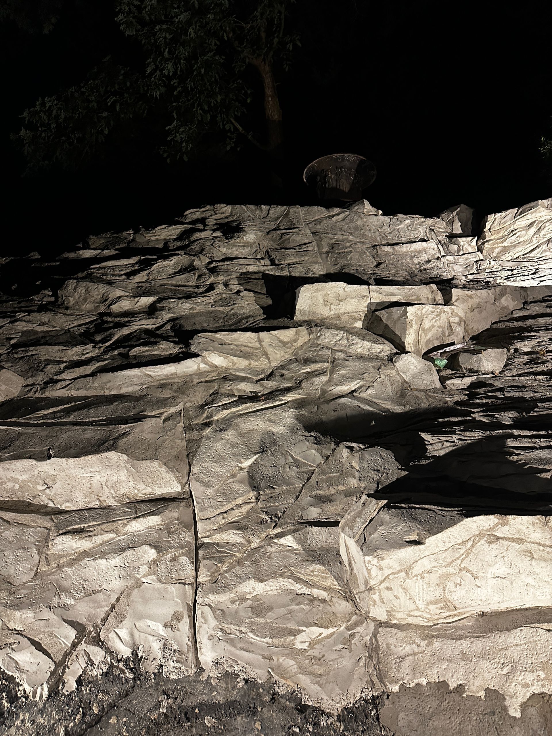 Stone wall with layered gray and white rocks, dark shadows, and a tree above.