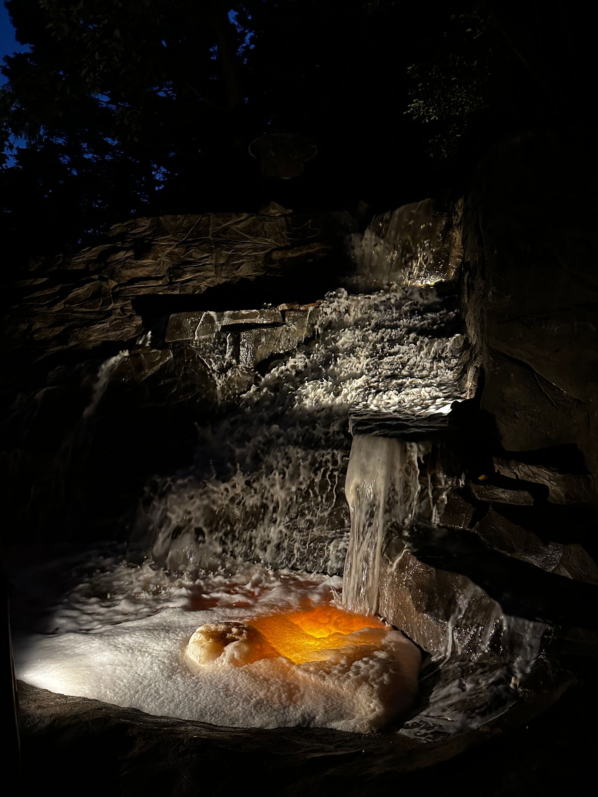 Waterfall at night, illuminated by spotlights, flowing into a pool with a golden-orange rock.