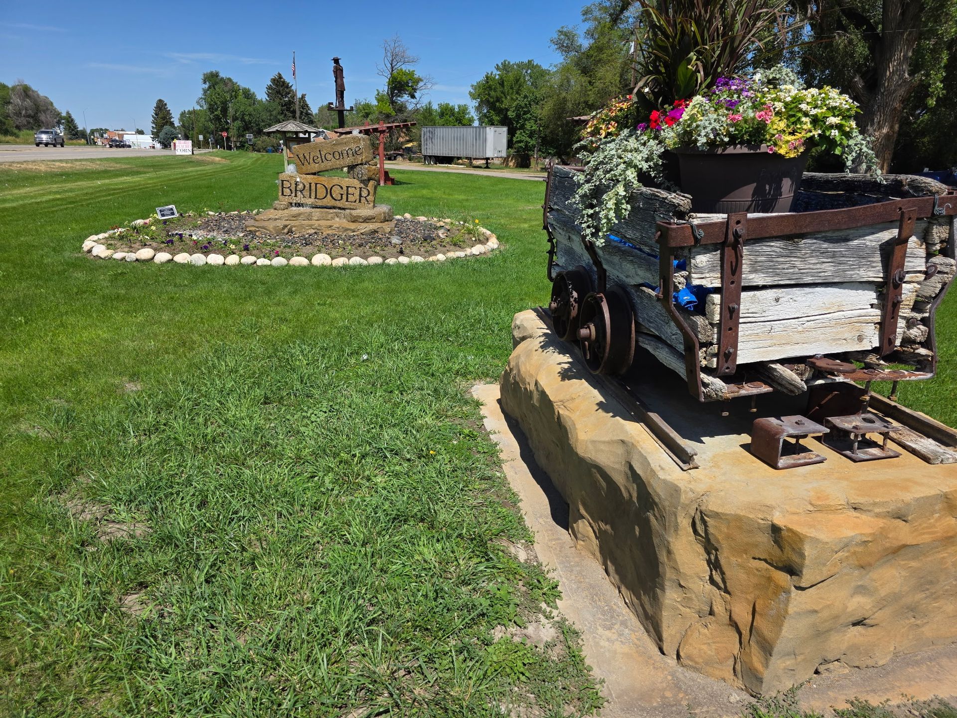 Antique mine cart filled with flowers on a stone base, green grass, another structure in the background.