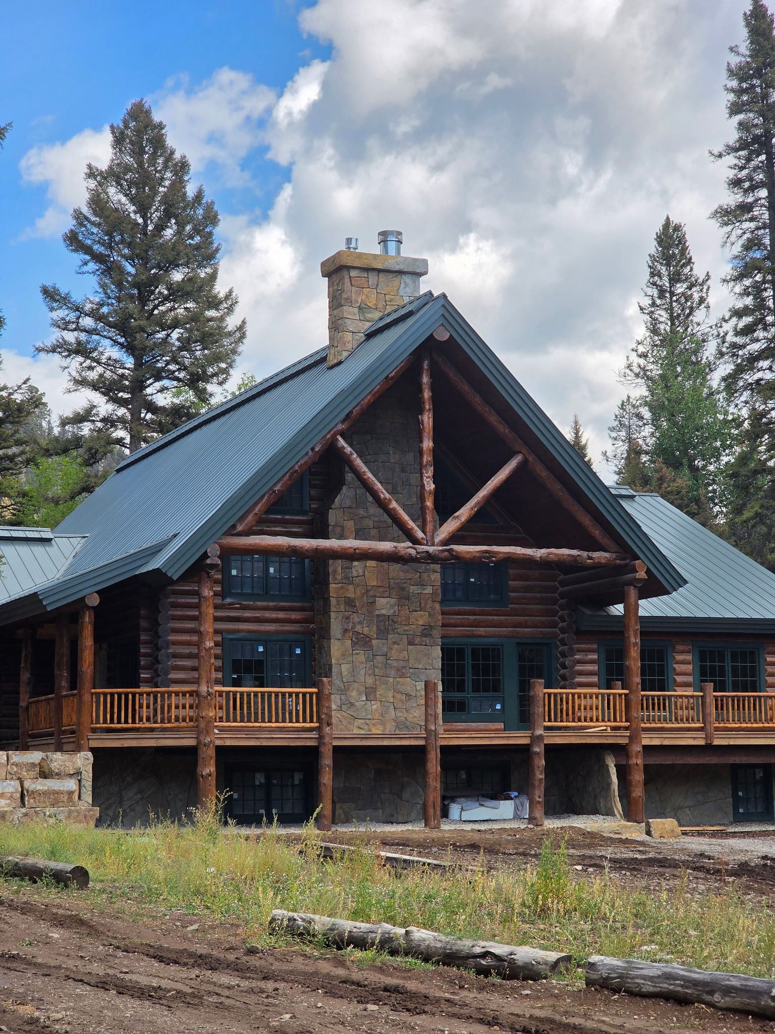 Log cabin with dark green metal roof, stone chimney, and wooden balconies in a wooded setting.