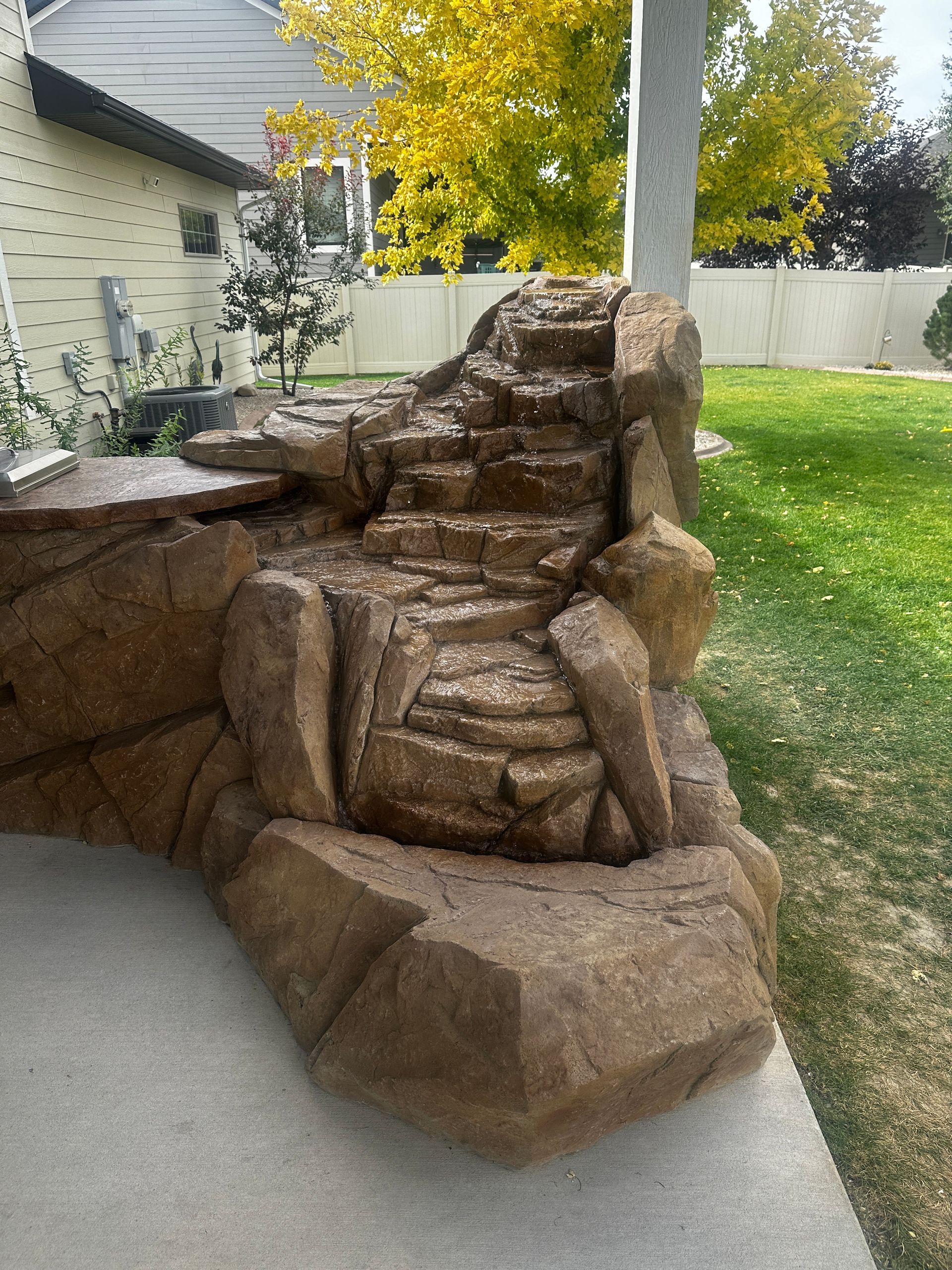 Rock waterfall fountain on a concrete patio in a yard with grass and fall foliage.