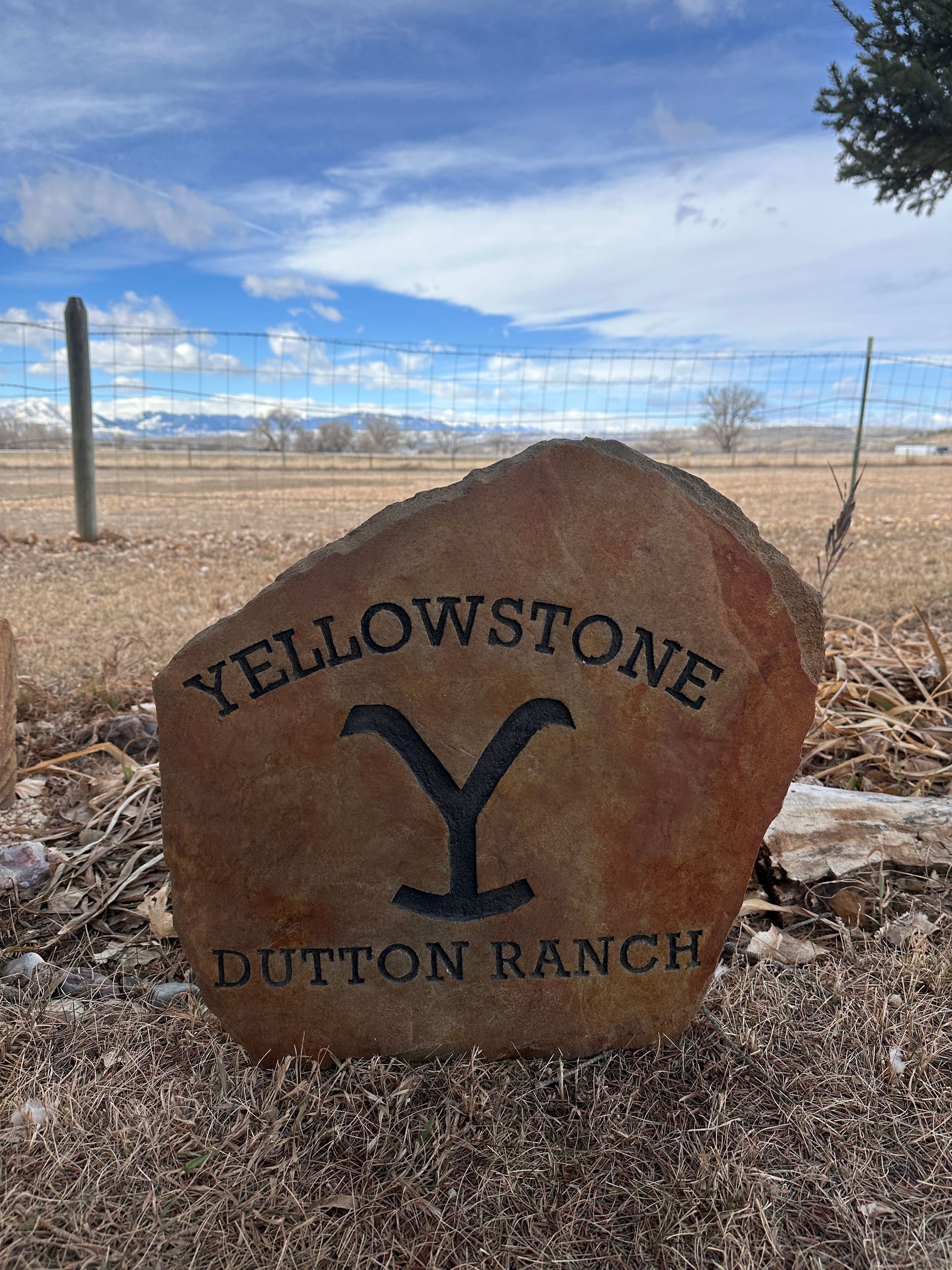 Yellowstone Dutton Ranch sign carved in stone against a rural backdrop; blue sky, mountains.