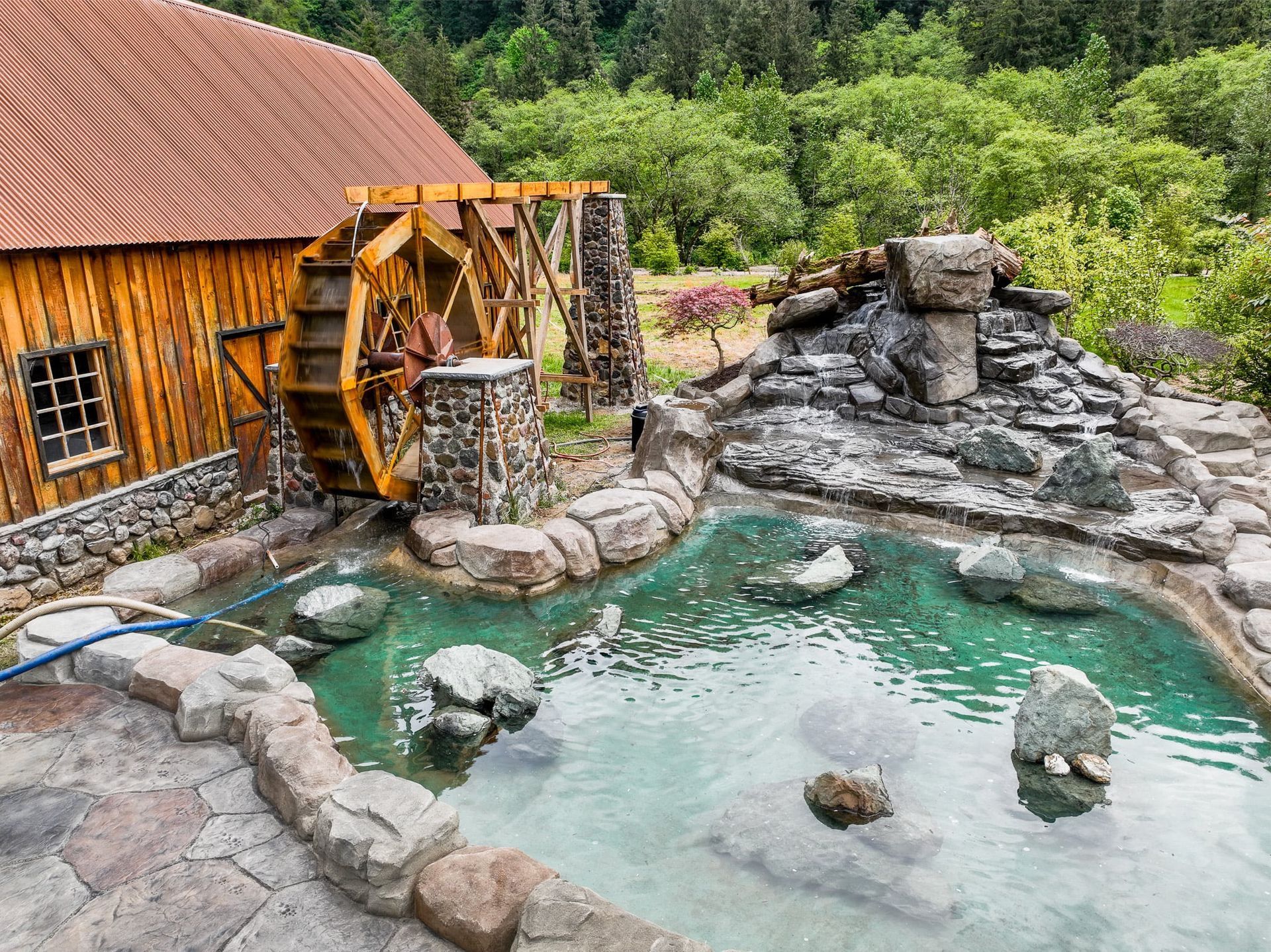 Wooden water wheel next to a stone-lined pool with clear turquoise water and a wooden building, surrounded by greenery.