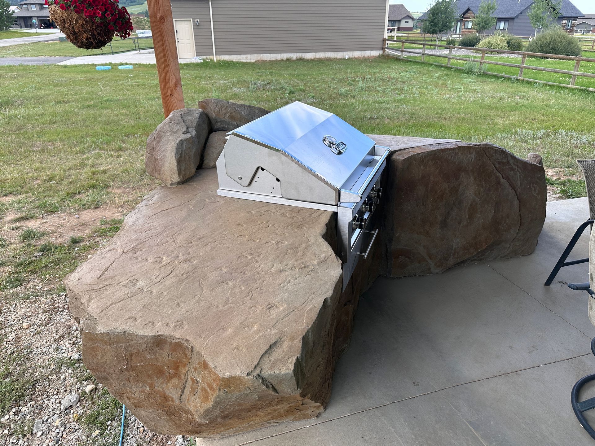 Outdoor barbecue grill built into large, brown boulders on a patio with grass and a hanging flower basket.