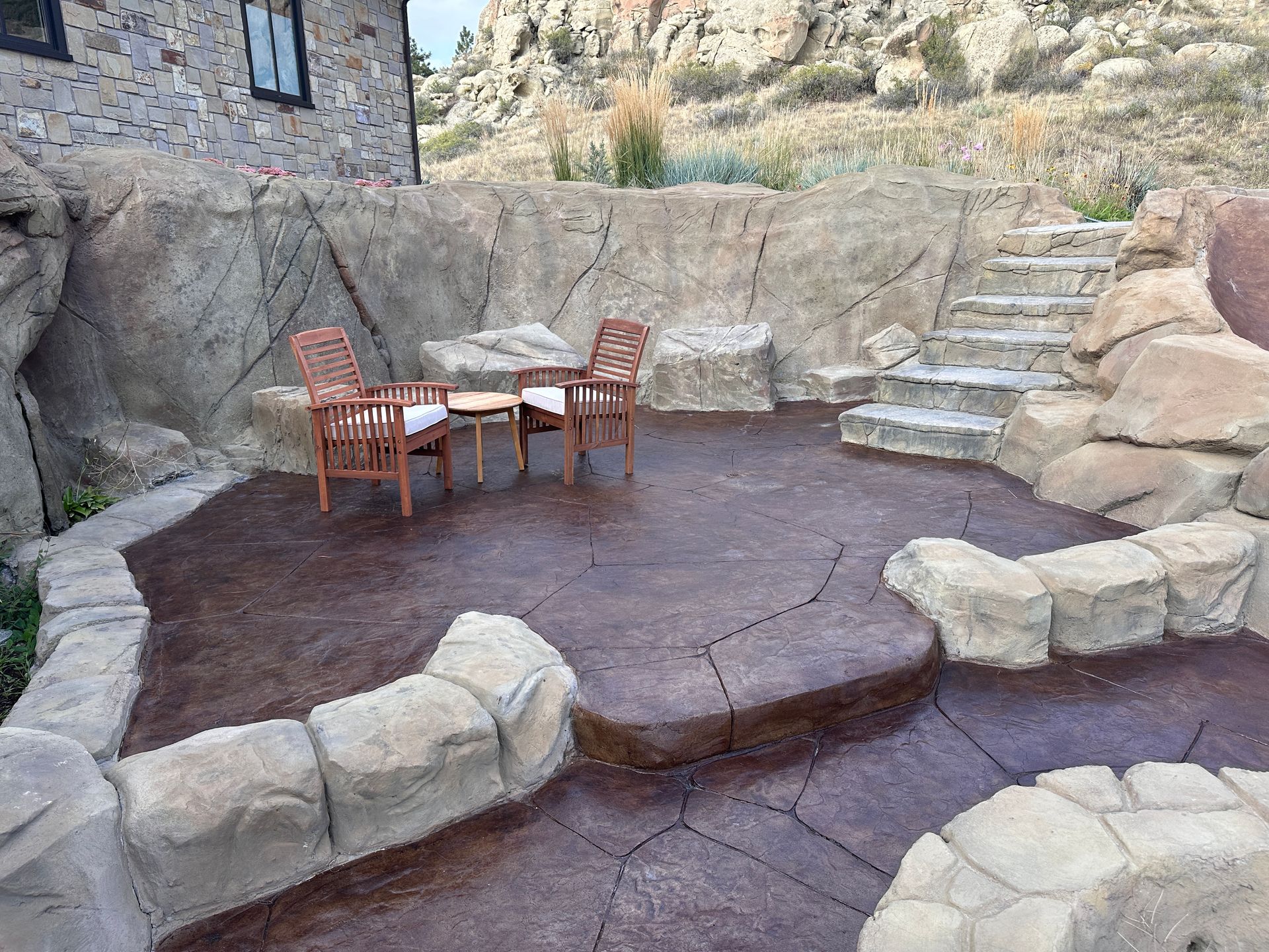 Brown patio with stone features and steps; two chairs and a table sit on the patio.
