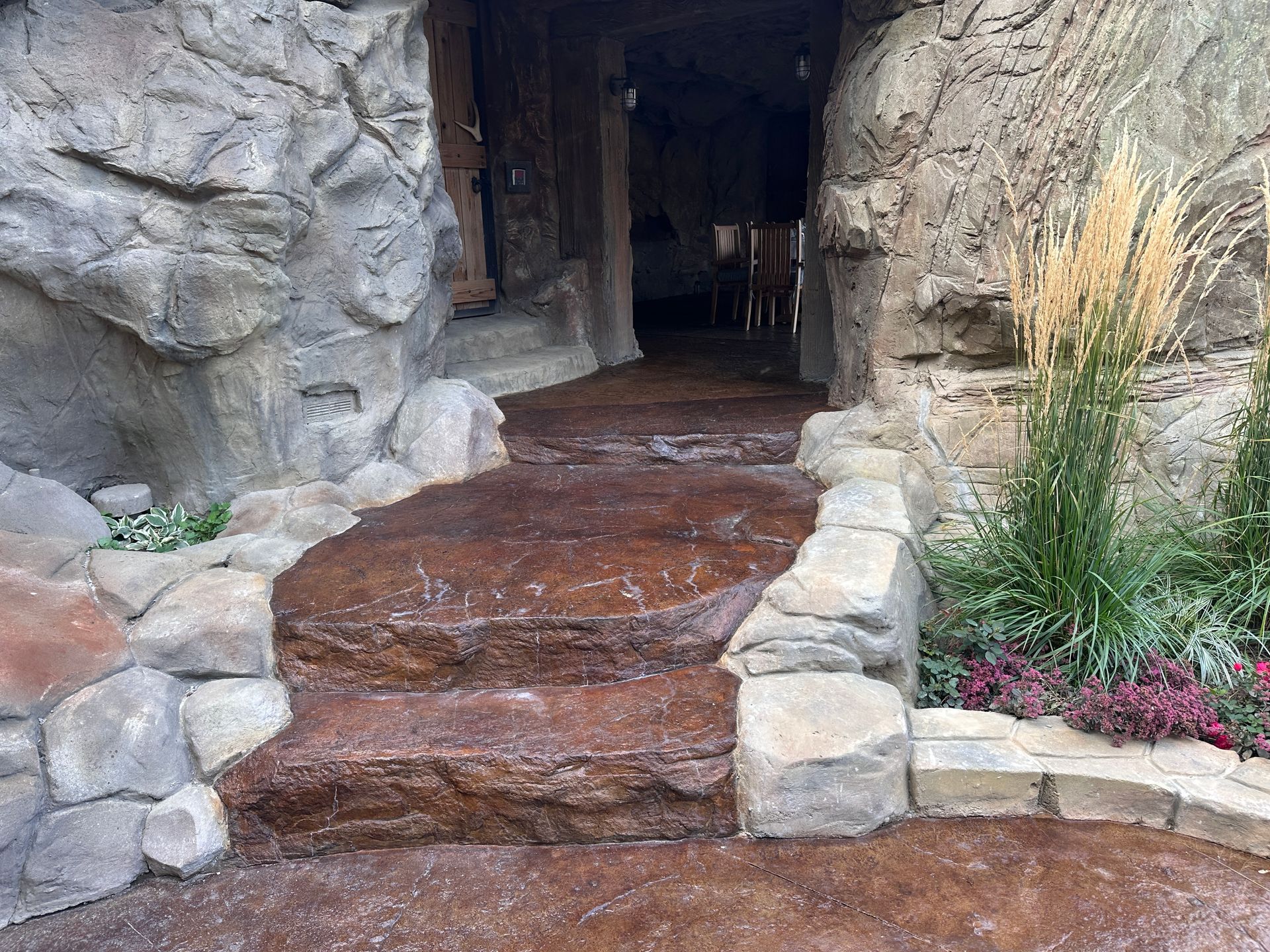 Stone steps leading into a cave-like entrance. Brown stone steps, rock walls, and plants.